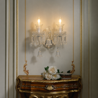 An ornate two-light crystal wall sconce mounted on a beige paneled wall above a carved gold console table with white flowers.