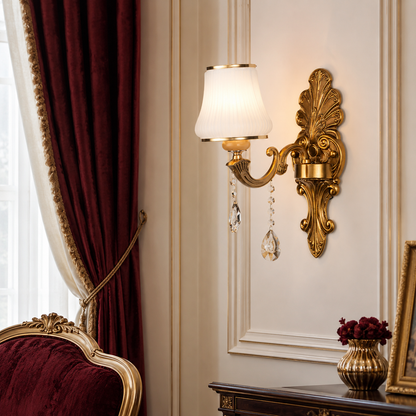 An ornate gold wall sconce with a white pleated shade and hanging crystal drops, mounted on a beige wall in a luxurious room with deep red curtains and a dark wood console table.