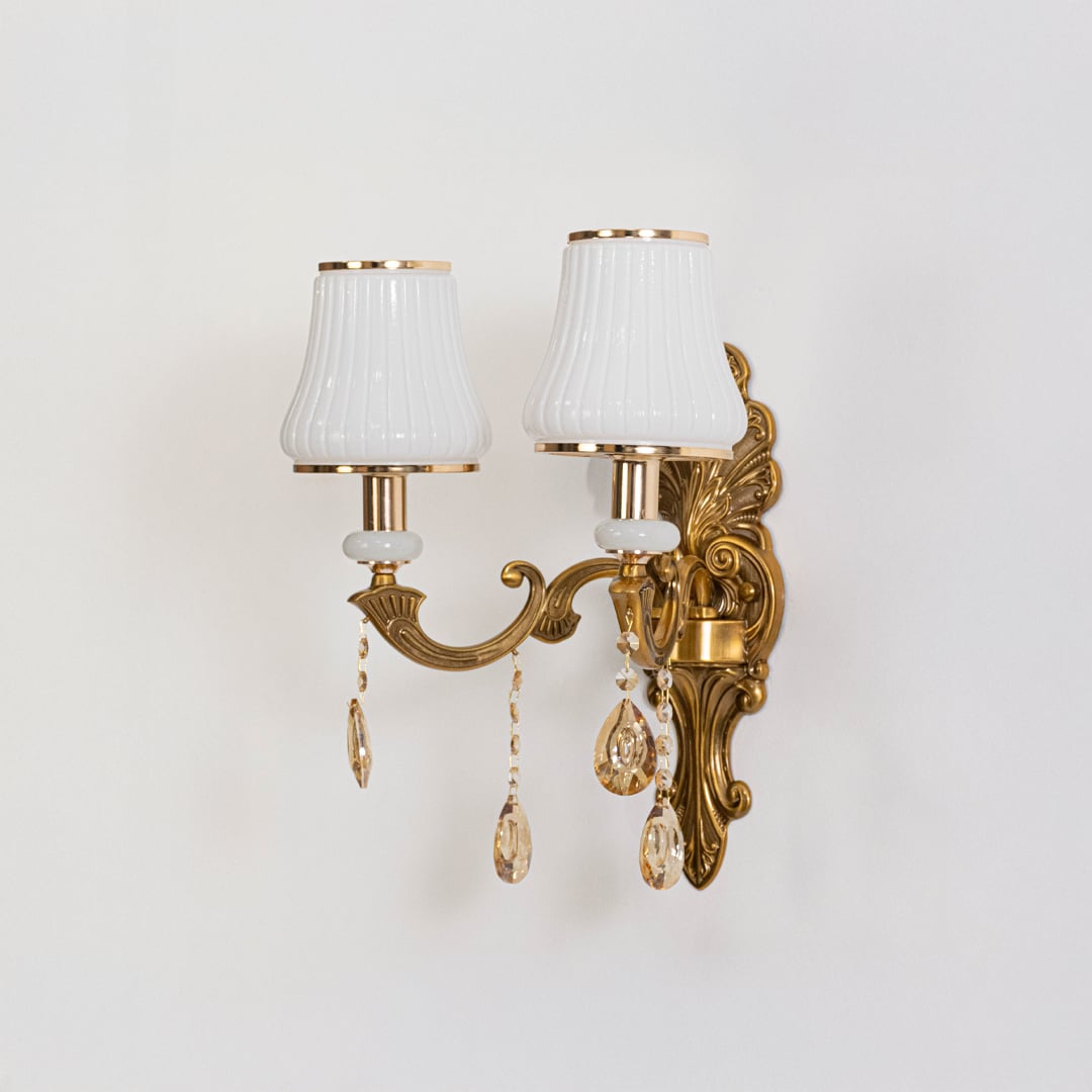 Two light crystal sconce with ornate gold base and white pleated shades shown switched off on white background.