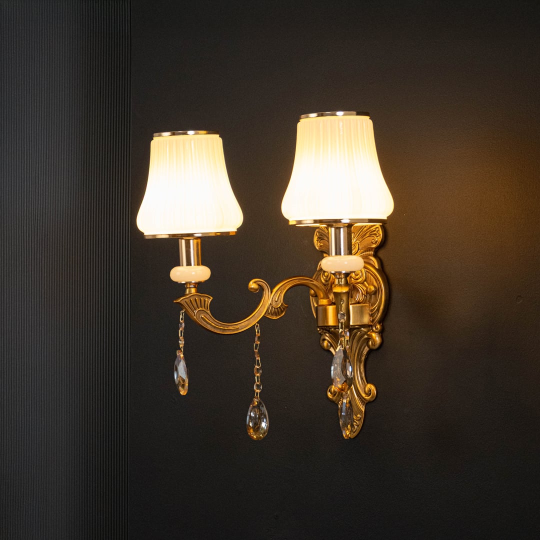 Luxury two light crystal sconce with ornate gold finish white pleated shades and crystal drops illuminated on black background.
