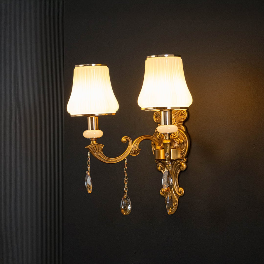Luxury two light crystal sconce with ornate gold finish white pleated shades and crystal drops illuminated on black background.