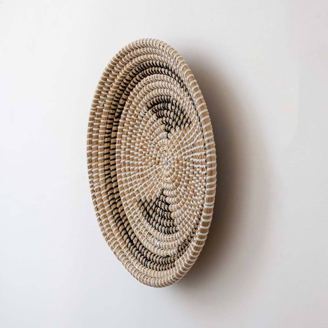 Handwoven seagrass round wall basket decor in natural beige with black crescent woven pattern shown from side on white background.