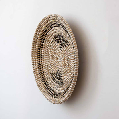 Handwoven seagrass round wall basket decor in natural beige with black crescent woven pattern shown from side on white background.