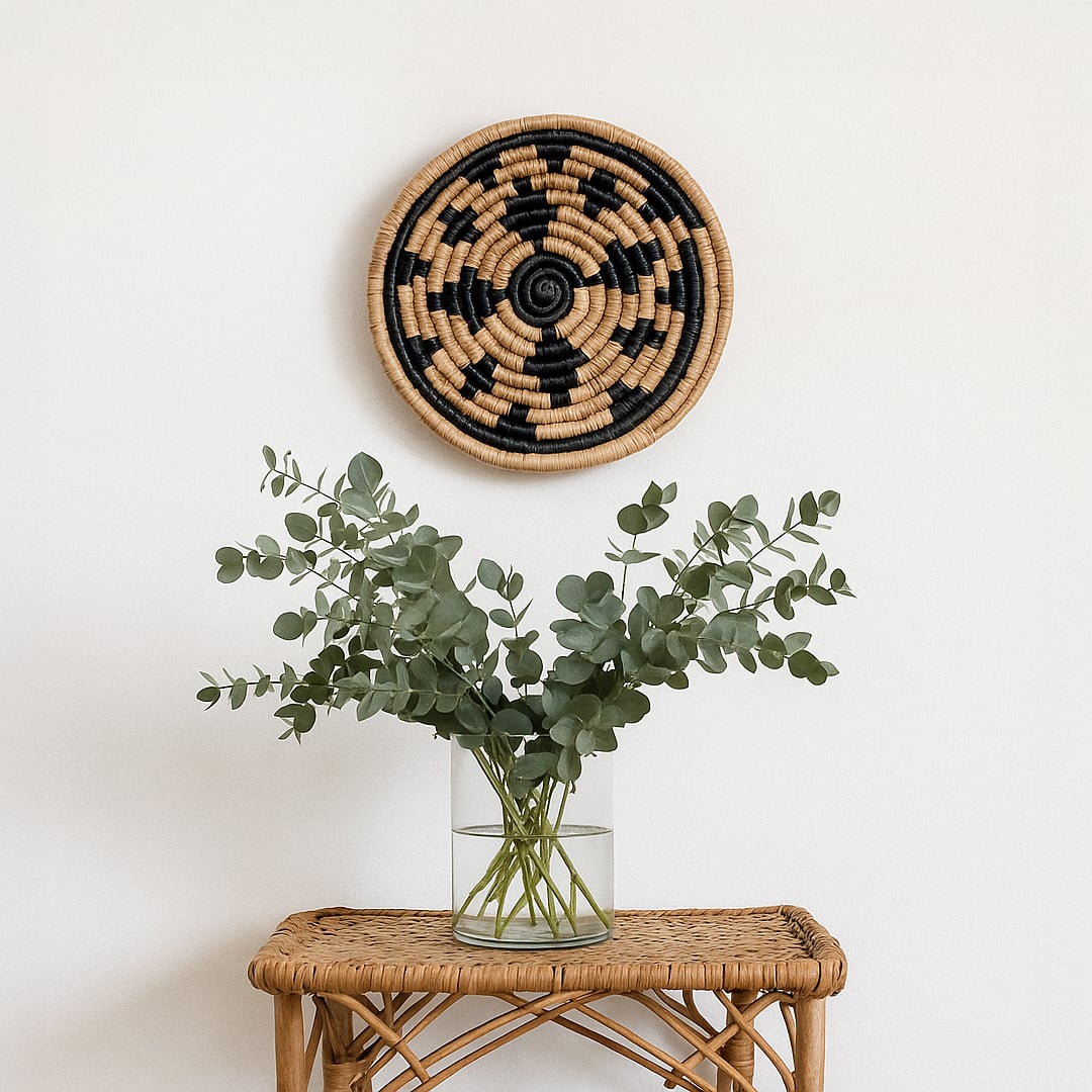 Main lifestyle image showing a handwoven round wall basket in natural and black tones styled above a rattan table with a glass vase and green foliage against a soft white wall background.