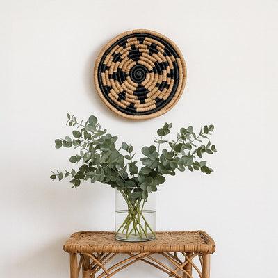 Main lifestyle image showing a handwoven round wall basket in natural and black tones styled above a rattan table with a glass vase and green foliage against a soft white wall background.