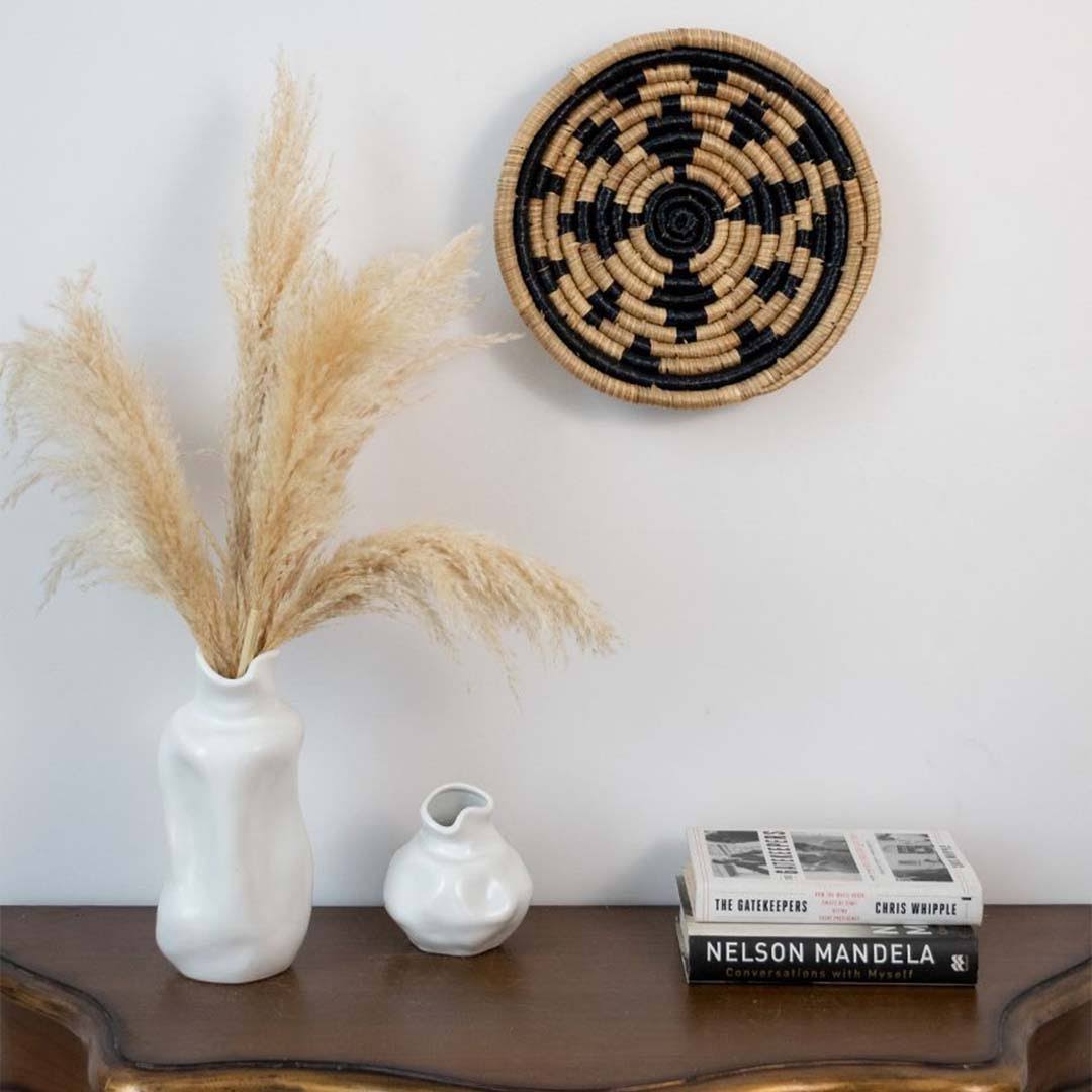 Decorative setup with a woven wall basket, pampas grass in a vase, and books on a wooden surface.