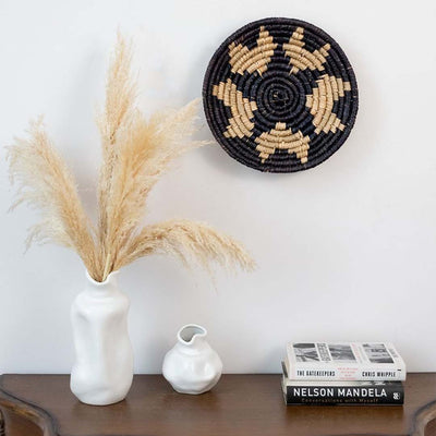 Decorative woven basket wall plate in neutral tones hanging on beige wall beside lamp and wooden side table.