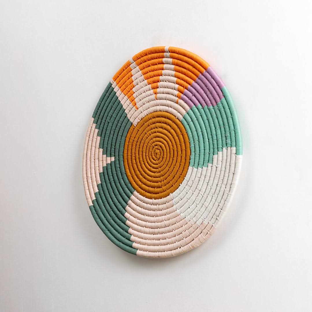 Round handcrafted multicolor seagrass wall basket with orange green cream and pink pattern on white background image.