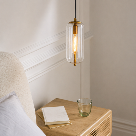 A modern pendant light with a clear glass cylinder and gold-toned metal accents, hanging above a wooden bedside table in a softly lit bedroom setting.