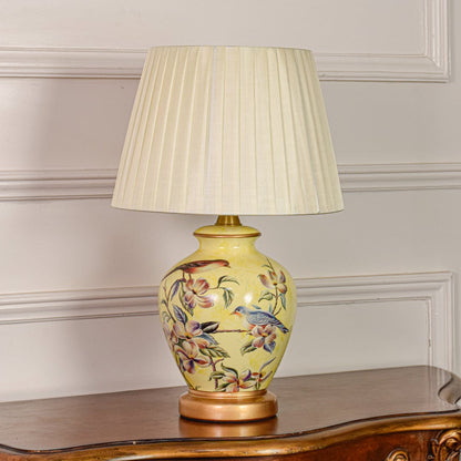Decorative yellow ceramic table lamp with floral bird design gold metal base and pleated fabric shade on off white backdrop.