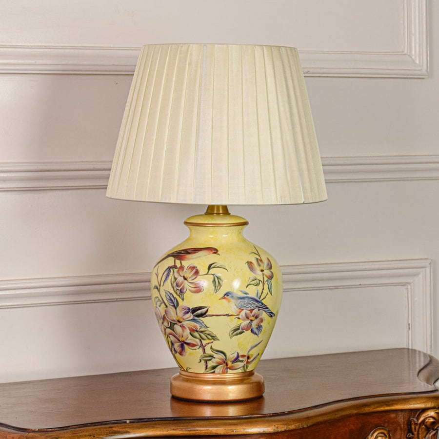 Decorative yellow ceramic table lamp with floral bird design gold metal base and pleated fabric shade on off white backdrop.