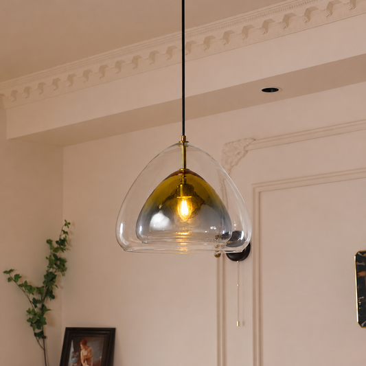 A modern pendant light with a clear glass dome shade and a reflective gold interior, suspended by a black cord in an elegant cream-colored room with decorative molding.