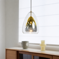 A modern pendant light with a clear teardrop-shaped glass shade and a brushed gold interior, hanging in a bright, minimalist room with a wooden sideboard and white window blinds.