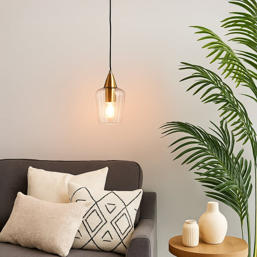 A clear glass pendant light with a gold top glowing warmly above a grey sofa and indoor plant creating a bright cozy living room ambiance.