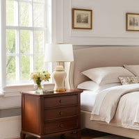 Wooden bedside table with ceramic table lamp and fabric shade placed beside bed adding warm ambient lighting to cozy bedroom decor.