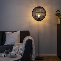Black floor lamp with woven rattan globe shade beside dark sofa in modern living room warm ambient lighting.