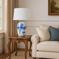 White ceramic table lamp with blue floral artwork and a soft white shade placed on a carved wooden side table adding a calm elegant touch to living room decor.