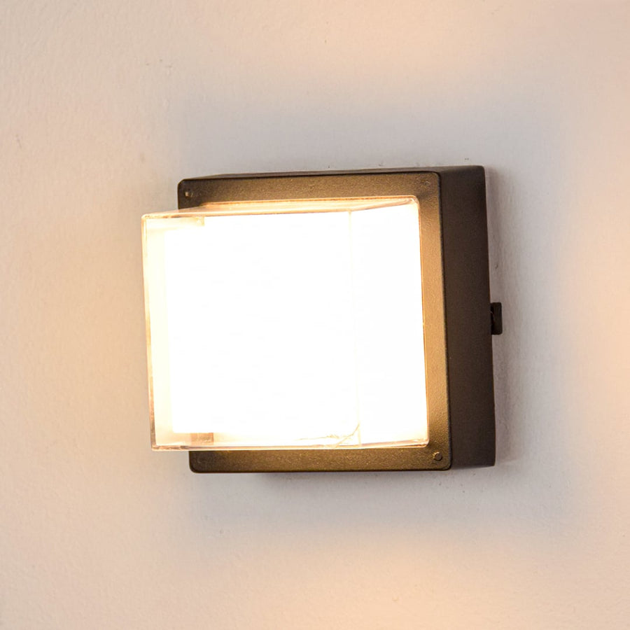 Black metal outdoor wall light switched on over white background spreading warm illumination through transparent glass cover.