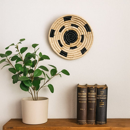 Lifestyle image of a woven round wall decor plate in natural and black tones styled above a wooden console with books and a potted plant in a calm modern interior.
