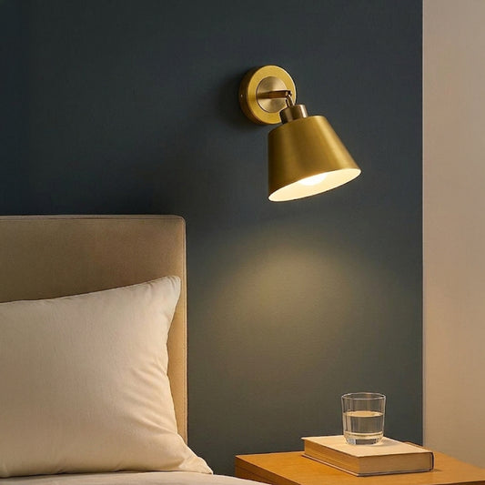 Gold metal wall light with cone shade casting warm glow styled beside bed creating cozy ambient lighting for bedroom decor.