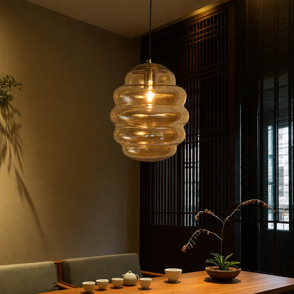 A modern pendant light with a ribbed, layered clear glass shade hanging over a wooden dining table in a warm, dimly lit interior.
