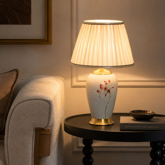 A table lamp with a pleated cream shade, a white ceramic base decorated with a red and green lotus motif, and gold-toned metal accents, styled on a dark round side table next to a beige armchair in a warmly lit room.