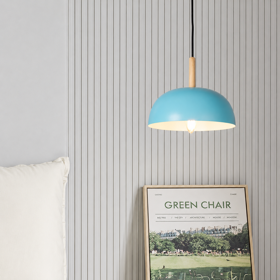 A modern pendant light with a light blue dome shade and a natural wood accent, hanging in a bedroom setting against a gray wood-paneled wall next to a white pillow and a framed 'Green Chair' poster.
