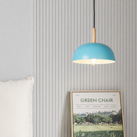 A modern pendant light with a light blue dome shade and a natural wood accent, hanging in a bedroom setting against a gray wood-paneled wall next to a white pillow and a framed 'Green Chair' poster.