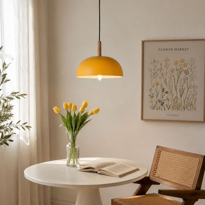 A warm yellow dome pendant light hangs over a small round white table in a softly lit room. A glass vase with yellow tulips, an open book, a woven chair, and a framed floral print are visible in the background.