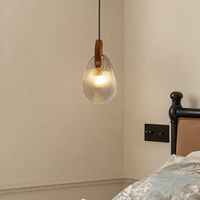 A modern pendant light with a ribbed clear glass shade, a brown leather strap, and a brass accent, hanging in a bedroom next to a bed with a black wooden headboard.