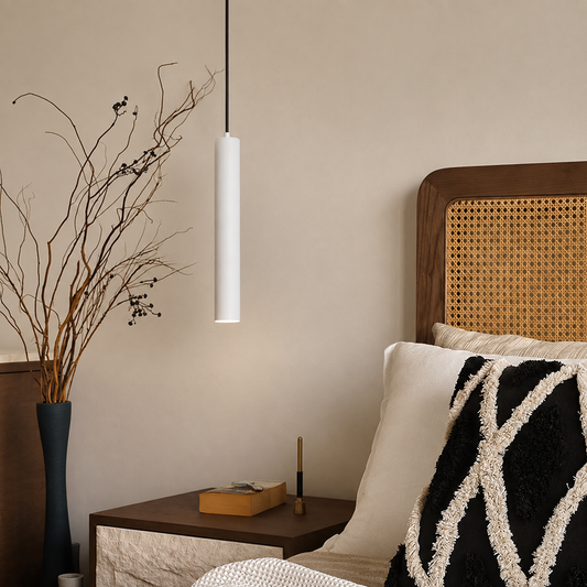 A minimalist white cylindrical pendant light hanging in a warm, modern bedroom setting next to a wooden nightstand and cane headboard.