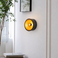 A modern wall-mounted sconce with a circular dark gray outer body and a glowing warm yellow-orange interior, installed on a white wall in a bright room with a window, plant, and framed art visible.