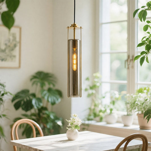 A modern cylindrical pendant light with a brushed brass metal frame and a warm glowing Edison bulb, hanging over a dining table in a bright, plant-filled room.