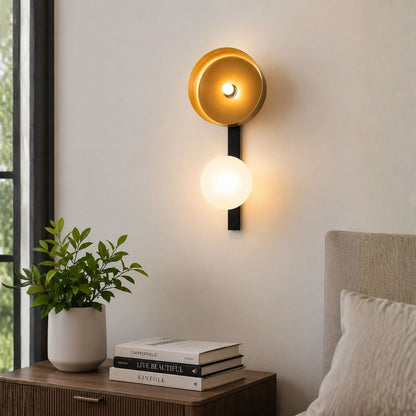 A modern wall-mounted sconce with a gold-toned upper shade and a glowing white lower globe, installed on a light wall above a bedside table with books and a plant.