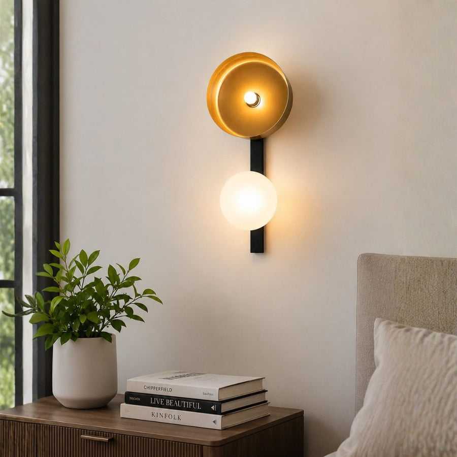 A modern wall-mounted sconce with a gold-toned upper shade and a glowing white lower globe, installed on a light wall above a bedside table with books and a plant.