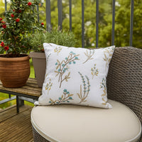 A white embroidered floral cushion placed on a wicker outdoor chair surrounded by green plants creating a fresh cozy balcony decor accent with soft natural lighting.
