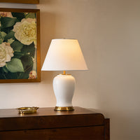 White fabric shade table lamp with ceramic white base casting warm ambient light on wooden console in luxury interior.