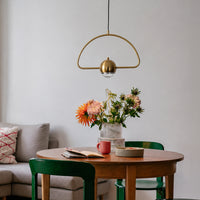A modern brass pendant light with a semi-circular frame and a dome-shaped bulb holder hangs over a round wooden dining table in a bright, contemporary room. The table is styled with a vase of colorful flowers, a red mug, and books, surrounded by green chairs.