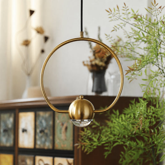 A modern brass pendant light with a circular ring and a spherical globe shade, hanging in a styled interior setting with a wooden cabinet and green plants in the background.