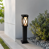 A black outdoor solar post lantern light with a warm glowing light, set on a stone ledge next to a white wall and green shrubs.