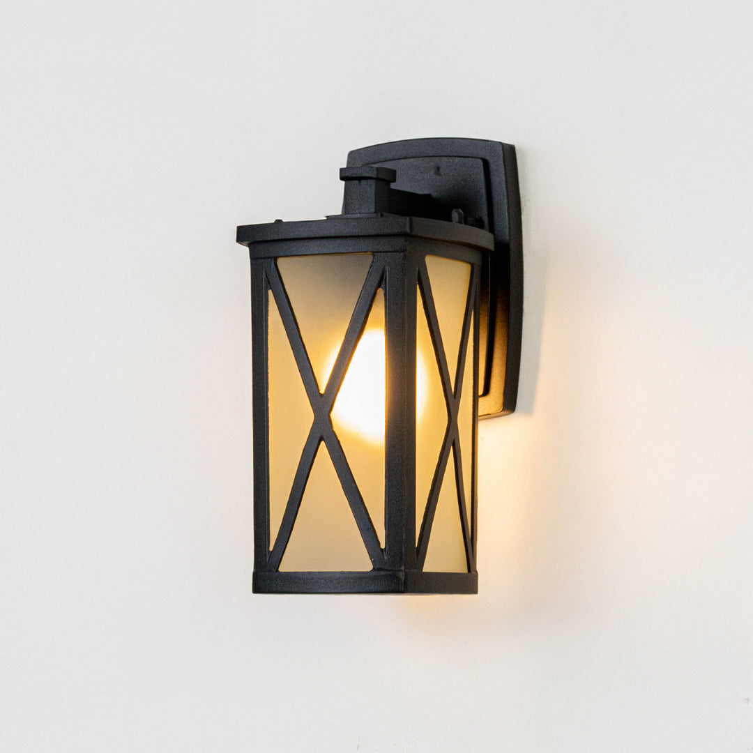 Black metal outdoor wall lantern on white background with frosted glass and X frame overlay casting a warm porch glow.