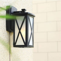 Black metal outdoor wall light lifestyle image featuring an X brace frosted glass shade mounted on a stone porch pillar.