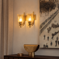 A two-light brass wall sconce with textured amber glass shades, mounted on a beige wall above a dark wooden console table with a decorative bowl and a large framed art print.