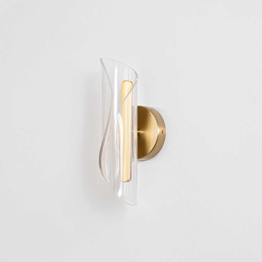 Gold wall light with sculpted clear glass shade shown on a bright white background with lights off for clarity.
