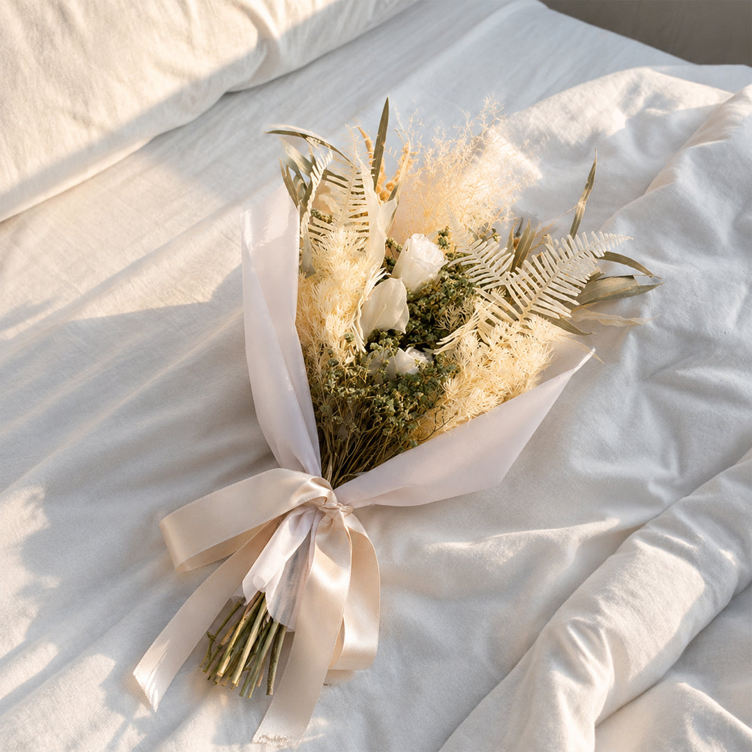 A minimalist cream beige dried flower bouquet wrapped in white paper resting on a rumpled white bedsheet in soft sunlight.