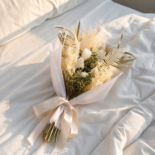 A minimalist cream beige dried flower bouquet wrapped in white paper resting on a rumpled white bedsheet in soft sunlight.