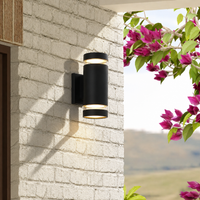 Modern black aluminum cylindrical dual up down wall sconce fixture on white brick house wall next to garden pink flowers.