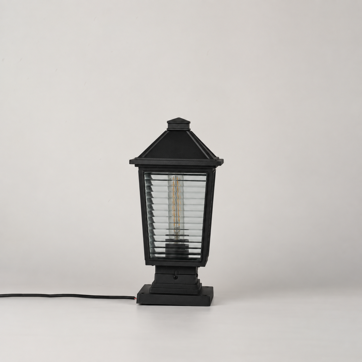 A black outdoor post cap light with a slatted glass shade and visible internal filament bulb, shown on a light gray studio background with its power cord extending to the left.