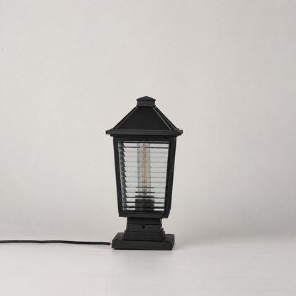 A black outdoor post cap light with a slatted glass shade and visible internal filament bulb, shown on a light gray studio background with its power cord extending to the left.