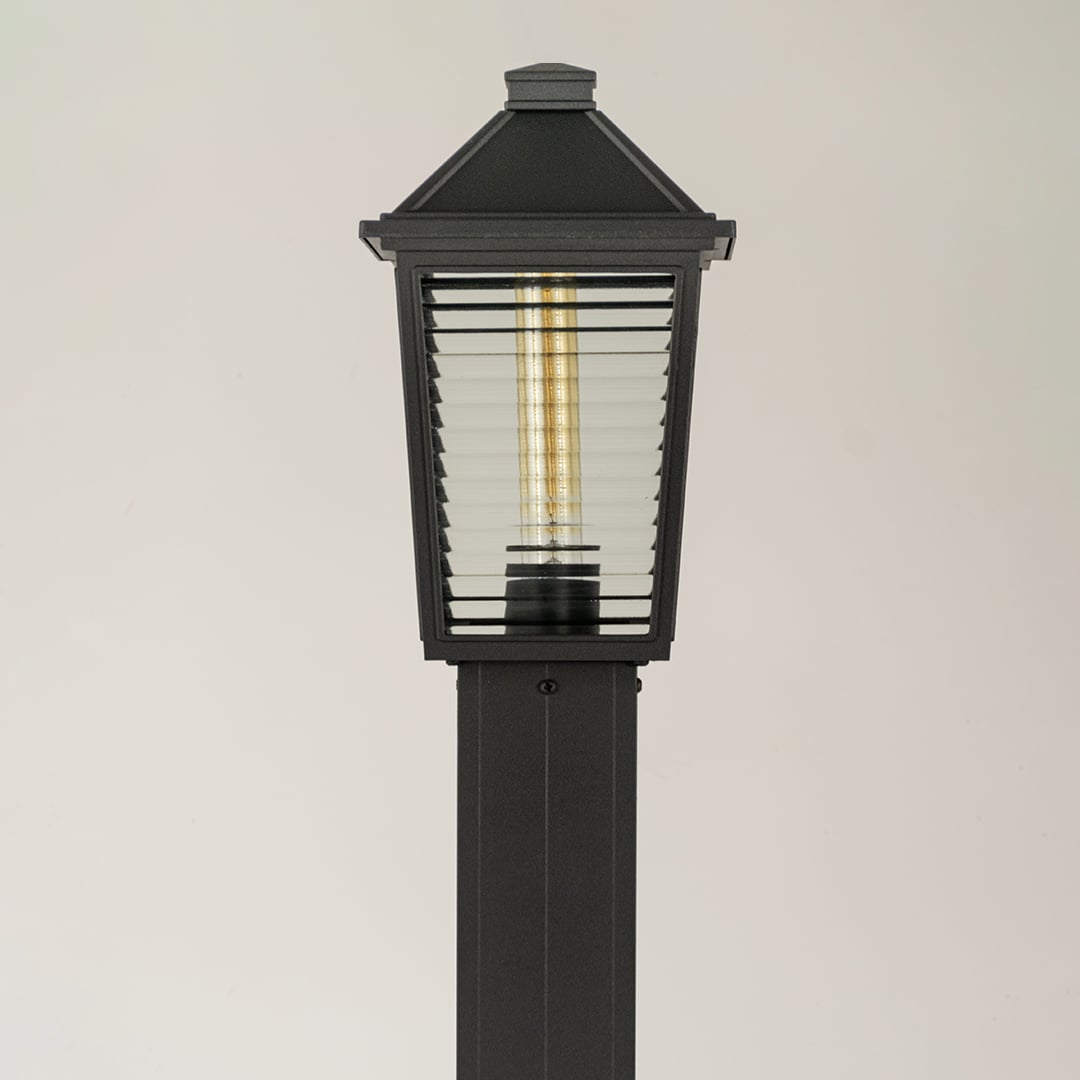 Closeup of a black outdoor pillar lamp showing warm light glowing through ribbed glass panels enhancing the classic lantern design and exterior detailing.